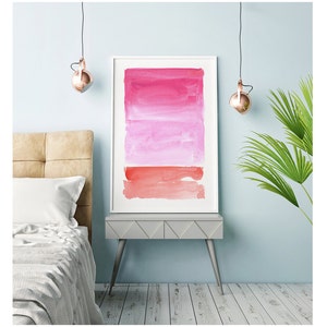 May include: Framed watercolor art print featuring a gradient of pink and red hues. The artwork is displayed in a white frame and rests on a gray side table. The room has a light blue wall, a bed, and a hanging light fixture.