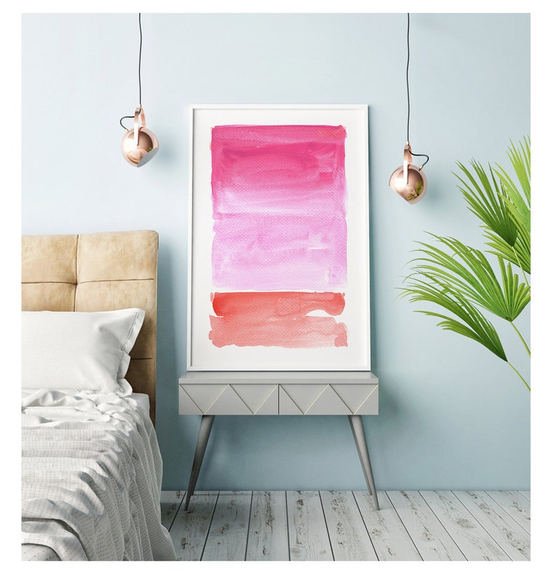 May include: Framed watercolor art print featuring a gradient of pink and red hues. The artwork is displayed in a white frame and rests on a gray side table. The room has a light blue wall, a bed, and a hanging light fixture.