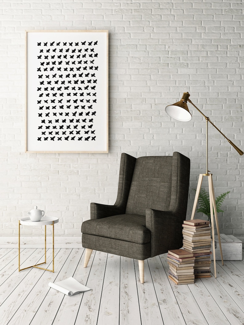 May include: A framed art print featuring a pattern of black butterfly shapes on a white background hangs on a white brick wall. A dark gray armchair with light wood legs sits nearby, with a small side table and a floor lamp.