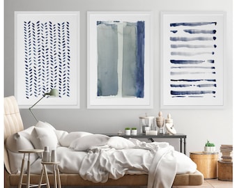 May include: Three framed abstract watercolor prints. The prints feature blue and gray watercolor designs. One print has a pattern of leaf-like shapes, another has vertical stripes, and the third has horizontal brushstrokes. The prints are displayed above a bed.
