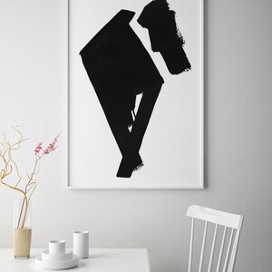 May include: Abstract black brushstroke art print in a white frame. The artwork features bold, geometric shapes and textures. The print is displayed above a white table with minimalist decor, including a vase and white dishes.