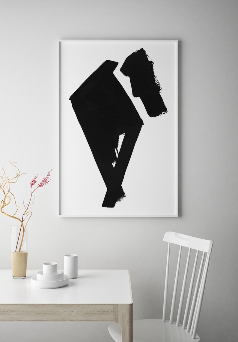 May include: Abstract black brushstroke art print in a white frame. The artwork features bold, geometric shapes and textures. The print is displayed above a white table with minimalist decor, including a vase and white dishes.