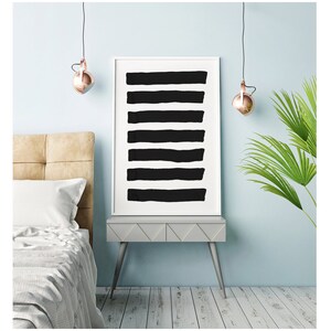 May include: A framed art print featuring eight thick, horizontal black brushstrokes on a white background. The artwork is displayed on a gray nightstand with silver legs, next to a bed with a beige headboard and a palm plant.