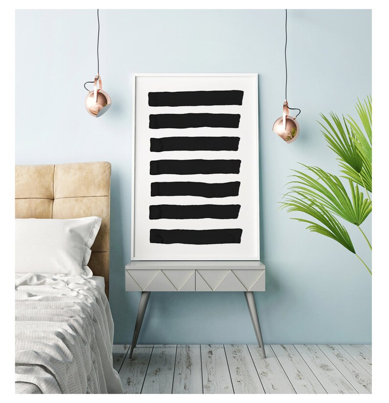 May include: A framed art print featuring eight thick, horizontal black brushstrokes on a white background. The artwork is displayed on a gray nightstand with silver legs, next to a bed with a beige headboard and a palm plant.