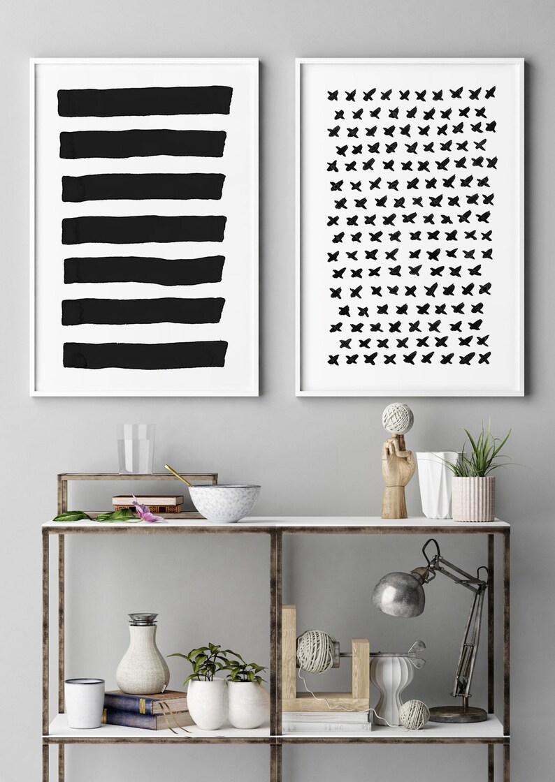 May include: Two framed abstract art prints. One features six thick, horizontal black brushstrokes on a white background. The other displays a pattern of small black butterfly shapes on a white background. The prints are displayed above a shelf with decorative items.