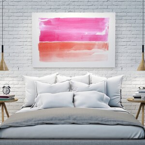 May include: A bedroom scene featuring a bed with white pillows and a gray comforter. Above the bed is a framed watercolor painting with pink and orange hues. Wooden bedside tables hold books, a clock, and a mug. Two pendant lights hang from the ceiling.