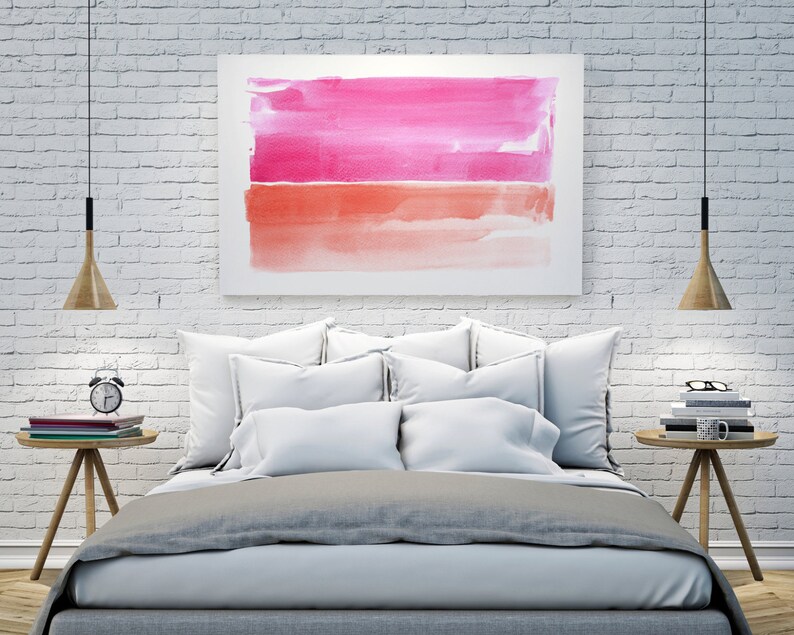 May include: A bedroom scene featuring a bed with white pillows and a gray comforter. Above the bed is a framed watercolor painting with pink and orange hues. Wooden bedside tables hold books, a clock, and a mug. Two pendant lights hang from the ceiling.