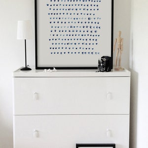 May include: A white dresser with three drawers, topped with a framed print of blue heart shapes. A small lamp, a vintage camera, and a wooden artist's mannequin are on the dresser. A framed print with text rests on the floor.