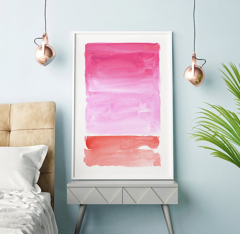 May include: A framed abstract watercolor painting with a white border. The artwork features a gradient of pink and red hues, creating a layered effect. The painting is displayed on a gray table with a light blue wall in the background.