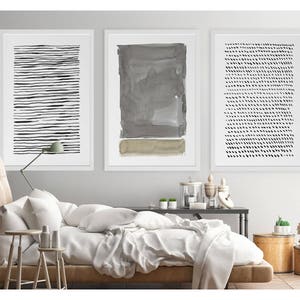 May include: Three framed abstract art prints. The prints feature black and white line and dot patterns, and a gray watercolor design. The artwork is displayed above a bed with white bedding and a wooden side table.