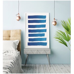 May include: A framed art print featuring eight horizontal blue brushstrokes against a white background. The artwork is displayed on a gray nightstand with silver legs, next to a bed with a beige headboard and a palm plant.