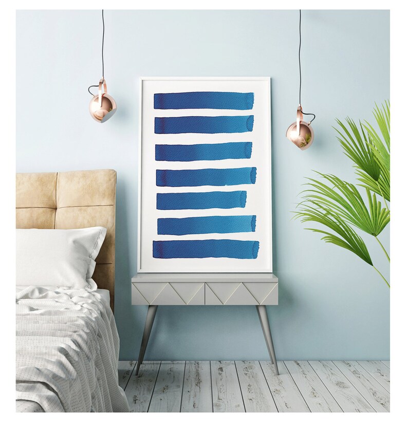 May include: A framed art print featuring eight horizontal blue brushstrokes against a white background. The artwork is displayed on a gray nightstand with silver legs, next to a bed with a beige headboard and a palm plant.