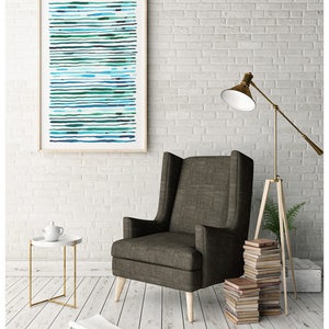 May include: An abstract art print in a wooden frame, featuring horizontal lines in shades of blue and green. The artwork hangs above a dark gray armchair with wooden legs. A white side table with a gold frame and a floor lamp complete the interior design.