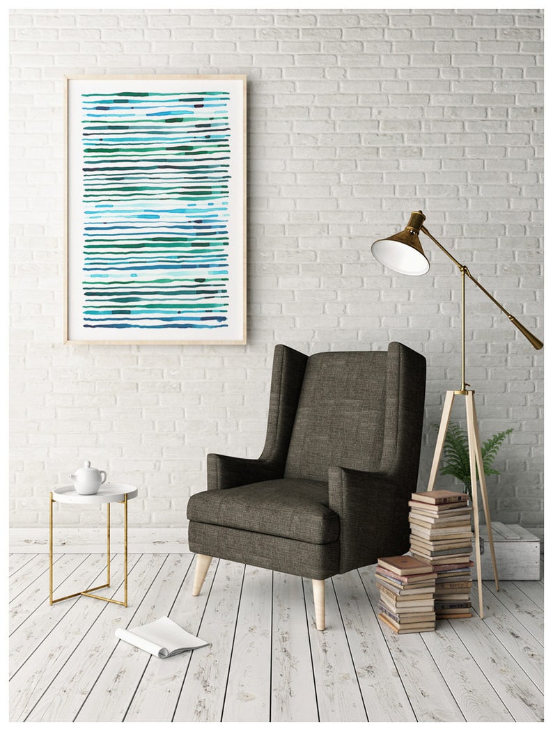 May include: An abstract art print in a wooden frame, featuring horizontal lines in shades of blue and green. The artwork hangs above a dark gray armchair with wooden legs. A white side table with a gold frame and a floor lamp complete the interior design.