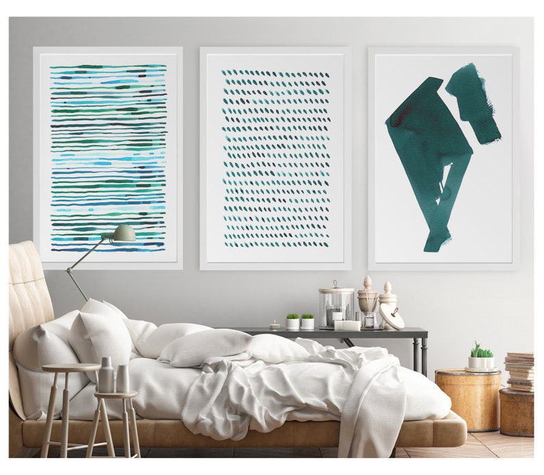 May include: Three framed abstract art prints. The prints feature watercolor designs in shades of teal and blue. One print has horizontal lines, another has diagonal dashes, and the third has geometric shapes. The prints are displayed above a bed with white bedding.