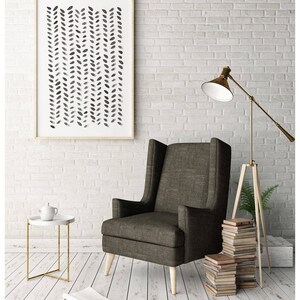 May include: A framed abstract art print with a pattern of black leaf shapes hangs above a dark gray armchair. A small white side table with a gold frame holds a teapot. A floor lamp and a stack of books complete the cozy interior.