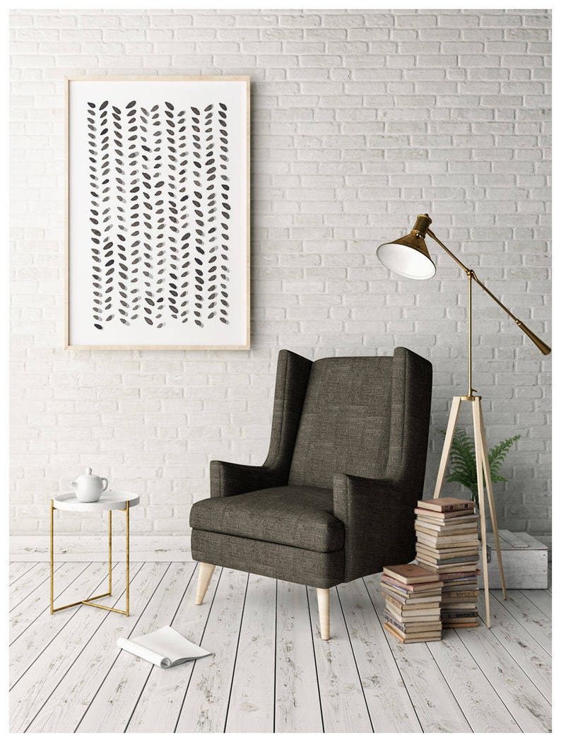 May include: A framed abstract art print with a pattern of black leaf shapes hangs above a dark gray armchair. A small white side table with a gold frame holds a teapot. A floor lamp and a stack of books complete the cozy interior.