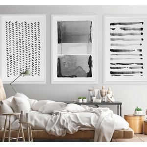 May include: Three framed abstract art prints in black and white. The prints feature watercolor designs: a leaf pattern, layered rectangles, and horizontal brushstrokes. The artwork is displayed above a bed with white bedding.