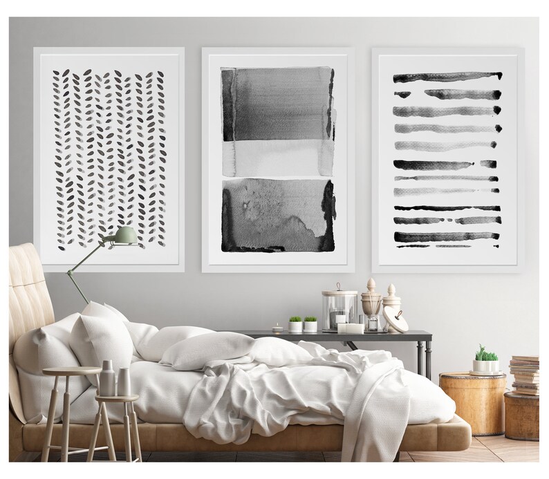 May include: Three framed abstract art prints in black and white. The prints feature watercolor designs: a leaf pattern, layered rectangles, and horizontal brushstrokes. The artwork is displayed above a bed with white bedding.