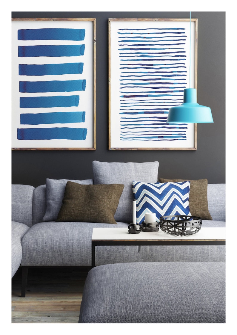 May include: Two framed abstract art prints with blue brushstrokes hang above a gray sofa with brown and blue pillows. A turquoise pendant lamp hangs from the ceiling. A white coffee table and a gray ottoman complete the living room setting.