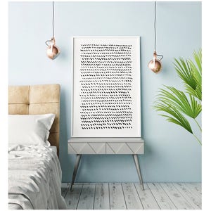 May include: A framed abstract art print with a white border, featuring a pattern of black dashes on a white background. The artwork is displayed on a gray side table with silver legs, next to a bed with a beige headboard. Copper-colored pendant lights hang above.