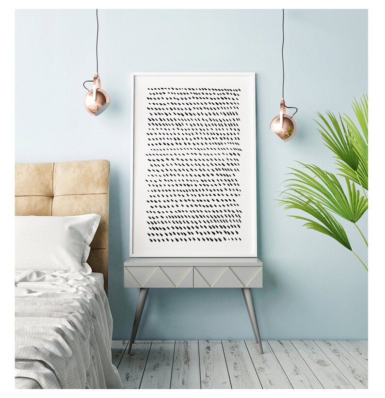 May include: A framed abstract art print with a white border, featuring a pattern of black dashes on a white background. The artwork is displayed on a gray side table with silver legs, next to a bed with a beige headboard. Copper-colored pendant lights hang above.
