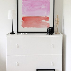 May include: A white dresser with three drawers, topped with a framed abstract watercolor painting in shades of pink and red. A small black lamp, a vintage camera, and a wooden artist's mannequin are also on the dresser. A framed print rests on the floor.