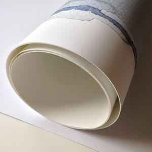 May include: Rolled watercolor paper with a blue and gray abstract design. The paper is white with a rolled edge, revealing the inside layers. The artwork features a watercolor painting with a blue river-like shape.