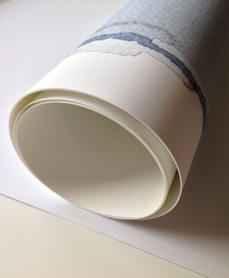 May include: Rolled watercolor paper with a blue and gray abstract design. The paper is white with a rolled edge, revealing the inside layers. The artwork features a watercolor painting with a blue river-like shape.
