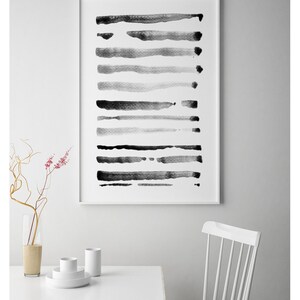 May include: Abstract art print featuring horizontal brushstrokes in varying shades of gray and black. The artwork is framed in a white border and hangs on a white wall. A white table and chair are in the foreground, with a vase of flowers.