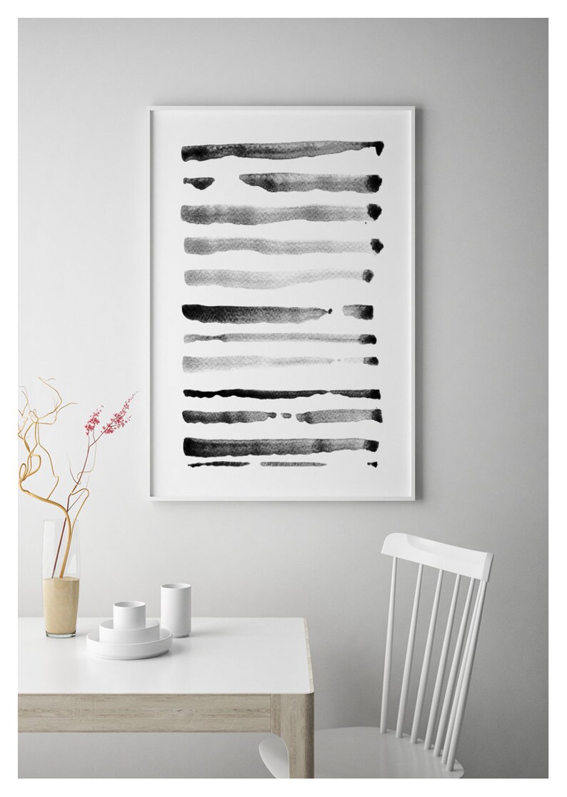 May include: Abstract art print featuring horizontal brushstrokes in varying shades of gray and black. The artwork is framed in a white border and hangs on a white wall. A white table and chair are in the foreground, with a vase of flowers.