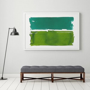 Op de afbeelding: Abstracte aquarel kunstdruk in een witte lijst. Het kunstwerk toont twee horizontale kleurvlakken: een turquoise groene bovenkant en een levendige groene onderkant. De print hangt boven een grijze bank met een donkerhouten frame.
