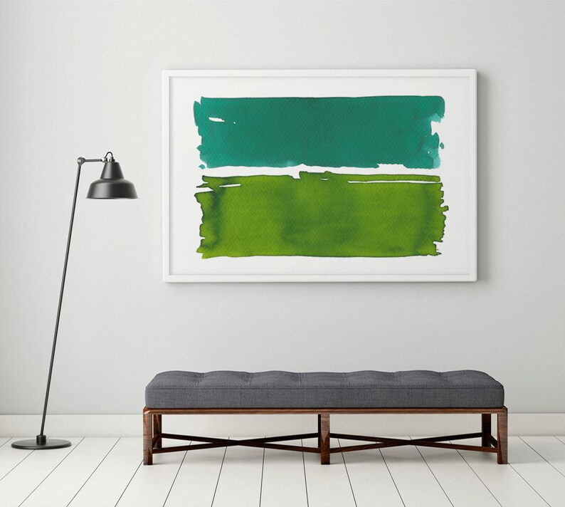 Op de afbeelding: Abstracte aquarel kunstdruk in een witte lijst. Het kunstwerk toont twee horizontale kleurvlakken: een turquoise groene bovenkant en een levendige groene onderkant. De print hangt boven een grijze bank met een donkerhouten frame.