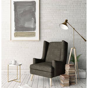 May include: A modern living room scene featuring a framed abstract watercolor painting in shades of gray and olive green. A tall, upholstered armchair sits next to a small side table with a teapot. A floor lamp and stacked books complete the decor.
