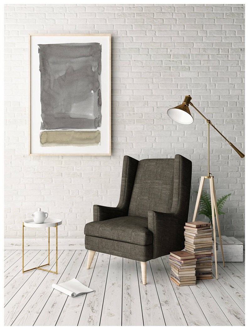 May include: A modern living room scene featuring a framed abstract watercolor painting in shades of gray and olive green. A tall, upholstered armchair sits next to a small side table with a teapot. A floor lamp and stacked books complete the decor.
