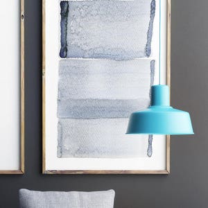 以下が含まれることがあります： 淡い青とグレーの抽象的なデザインの額入り水彩画。ターコイズ色のペンダントランプが天井から吊り下げられています。絵は木製のフレームに入っています。グレーと茶色のクッションが前景にあります。