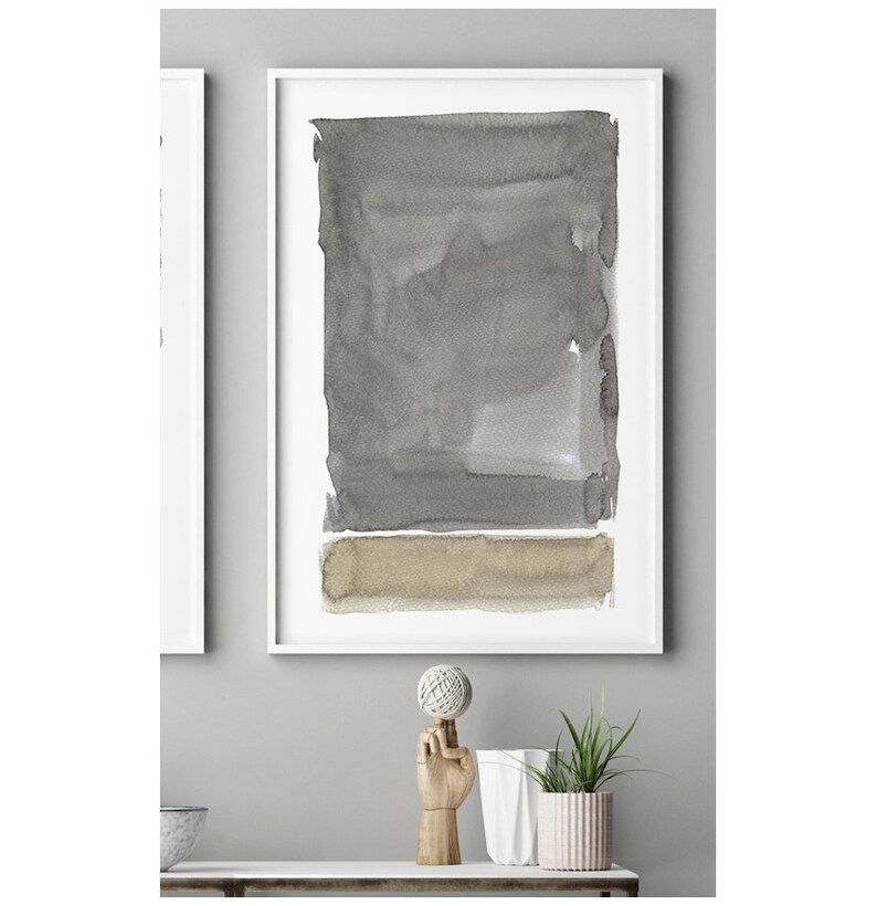 May include: Abstract watercolor art print in a white frame. The artwork features a large gray rectangular shape above a smaller, horizontal tan shape. The watercolor effect gives a soft, textured appearance. The art print is displayed on a white shelf.