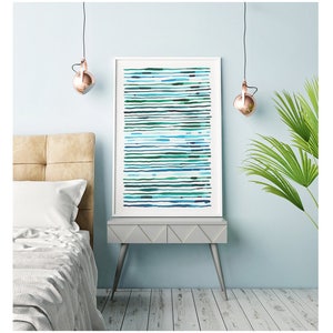 May include: A framed abstract art print featuring horizontal lines in shades of blue and green. The artwork is displayed on a gray side table with silver legs, next to a bed with a beige headboard. The room has a light blue wall and a copper-colored pendant light.