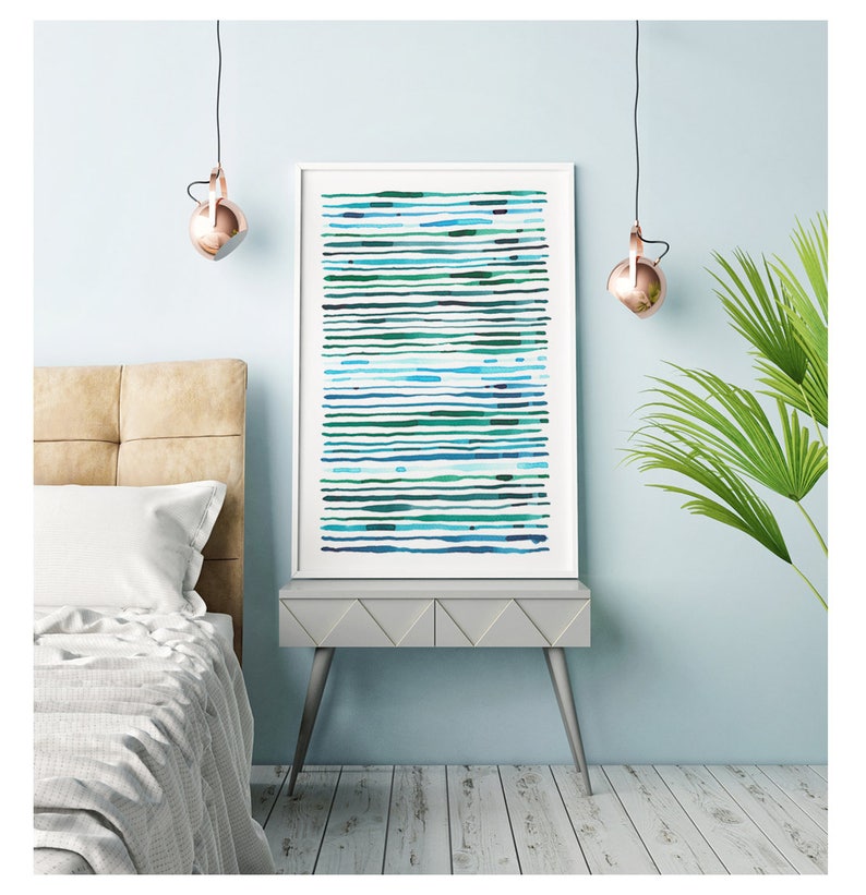 May include: A framed abstract art print featuring horizontal lines in shades of blue and green. The artwork is displayed on a gray side table with silver legs, next to a bed with a beige headboard. The room has a light blue wall and a copper-colored pendant light.