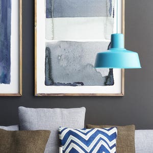 May include: A modern living room scene featuring abstract watercolor art in blue and gray tones, framed in wood. A turquoise pendant lamp hangs above a sofa with decorative pillows in blue, brown, and white. A coffee table holds candles and a black metal basket.