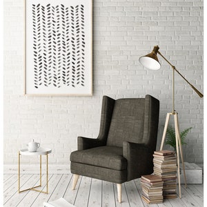 May include: A framed abstract art print with a neutral color palette hangs above a wingback chair. The print features a pattern of black and gray leaf-like shapes. A small side table with a white top and gold legs sits nearby.