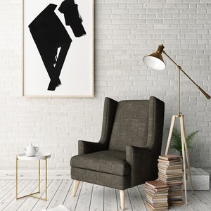 May include: An interior design scene featuring a tall, dark gray armchair with light wooden legs. A framed abstract black and white artwork hangs on a white brick wall. A small white side table with a gold frame and a floor lamp are also present.