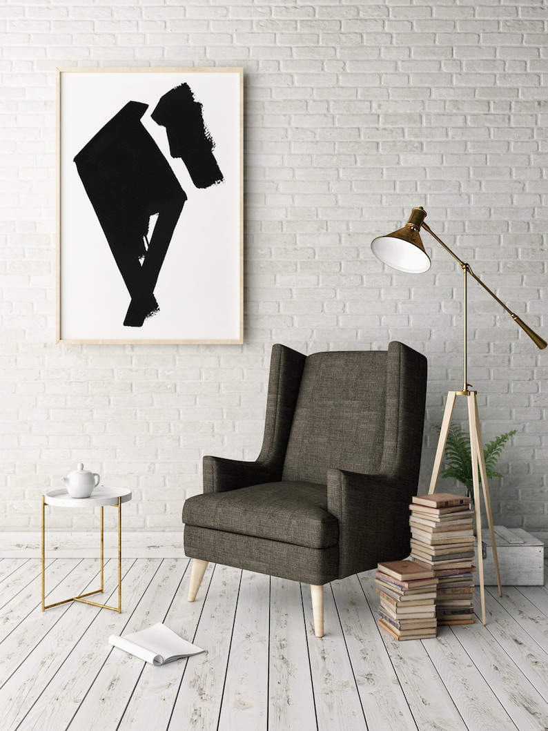 May include: An interior design scene featuring a tall, dark gray armchair with light wooden legs. A framed abstract black and white artwork hangs on a white brick wall. A small white side table with a gold frame and a floor lamp are also present.