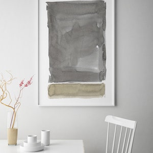 May include: Abstract watercolor art print in a white frame. The artwork features a rectangular composition with shades of gray and a tan horizontal band at the bottom. The print is displayed above a white table with minimalist decor.