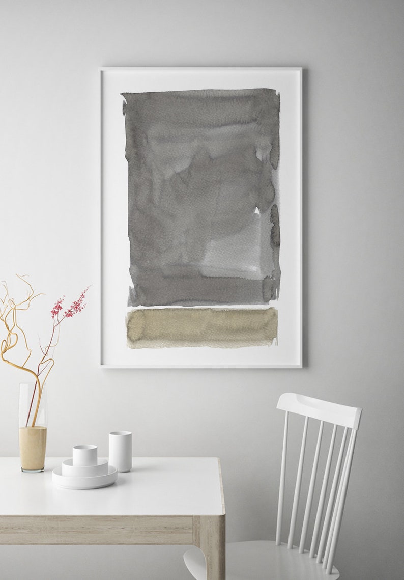 May include: Abstract watercolor art print in a white frame. The artwork features a rectangular composition with shades of gray and a tan horizontal band at the bottom. The print is displayed above a white table with minimalist decor.
