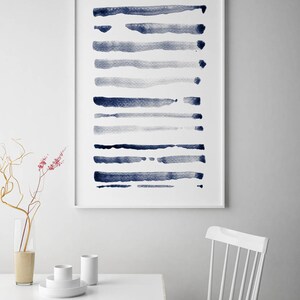 以下が含まれることがあります： 白い背景に水平な青い筆致が描かれた抽象的なアートプリント。白いフレームに入っており、白い壁に飾られています。この画像は、ミニマリスト的な美学を暗示しています。水彩画を思わせる作品です。