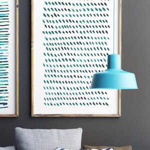 May include: Two framed abstract art prints with a wooden frame. One print features horizontal teal lines, the other has a pattern of teal brushstrokes. A turquoise pendant lamp hangs in front of the artwork. Gray and patterned pillows are in the foreground.
