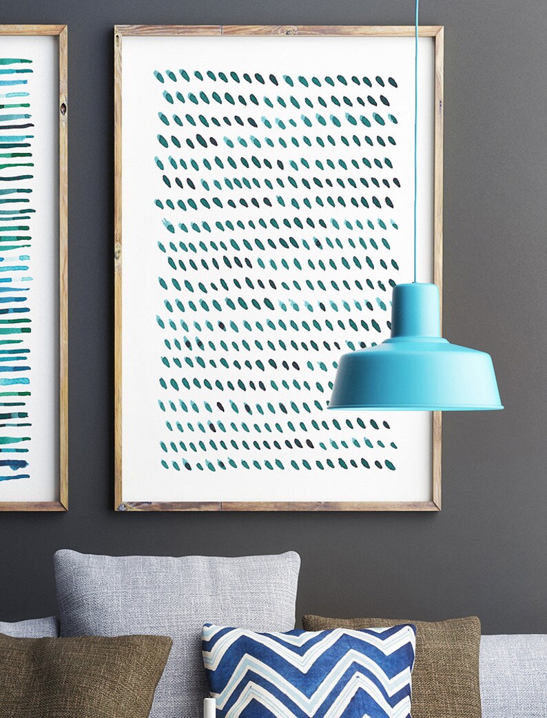 May include: Two framed abstract art prints with a wooden frame. One print features horizontal teal lines, the other has a pattern of teal brushstrokes. A turquoise pendant lamp hangs in front of the artwork. Gray and patterned pillows are in the foreground.