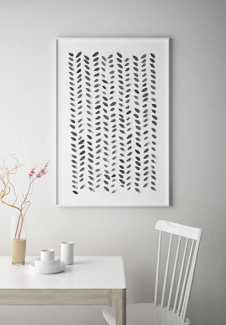 May include: A framed abstract art print featuring a pattern of dark gray leaf-like shapes arranged in vertical rows against a white background. The artwork is displayed on a white wall above a white table and chair.