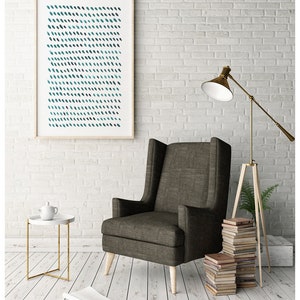 May include: A framed abstract art print with a pattern of teal and dark blue brushstrokes hangs on a white brick wall. A gray armchair with wooden legs sits below the artwork. A small side table and a floor lamp are also in the room.